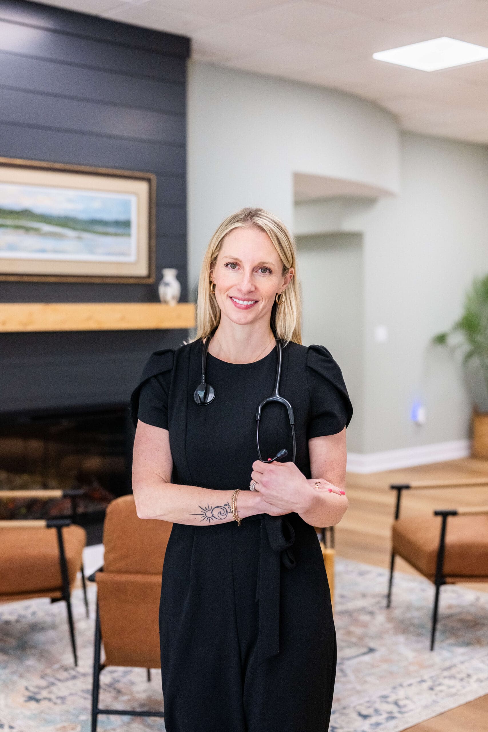 A woman in a black dress and a stethoscope stands in a modern waiting room with a fireplace