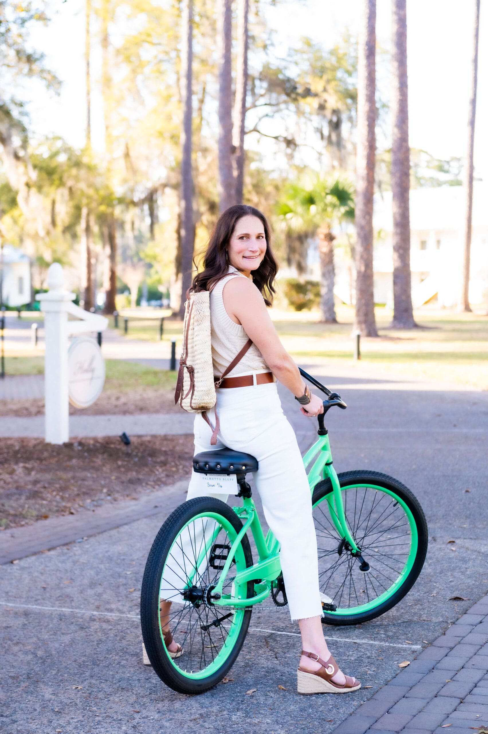 A woman smiles over her shoulder while standing over a green bicycle in white pants and tan blouse after finding what to wear for your branding photoshoot
