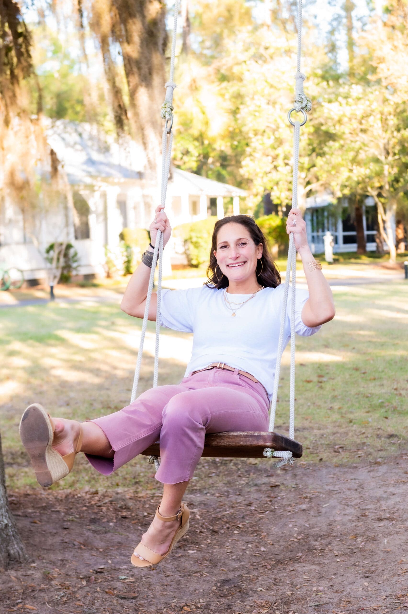 A woman sits smiling while swinging on a wooden tree swing in pink pants after discovering what to wear for your branding photoshoot