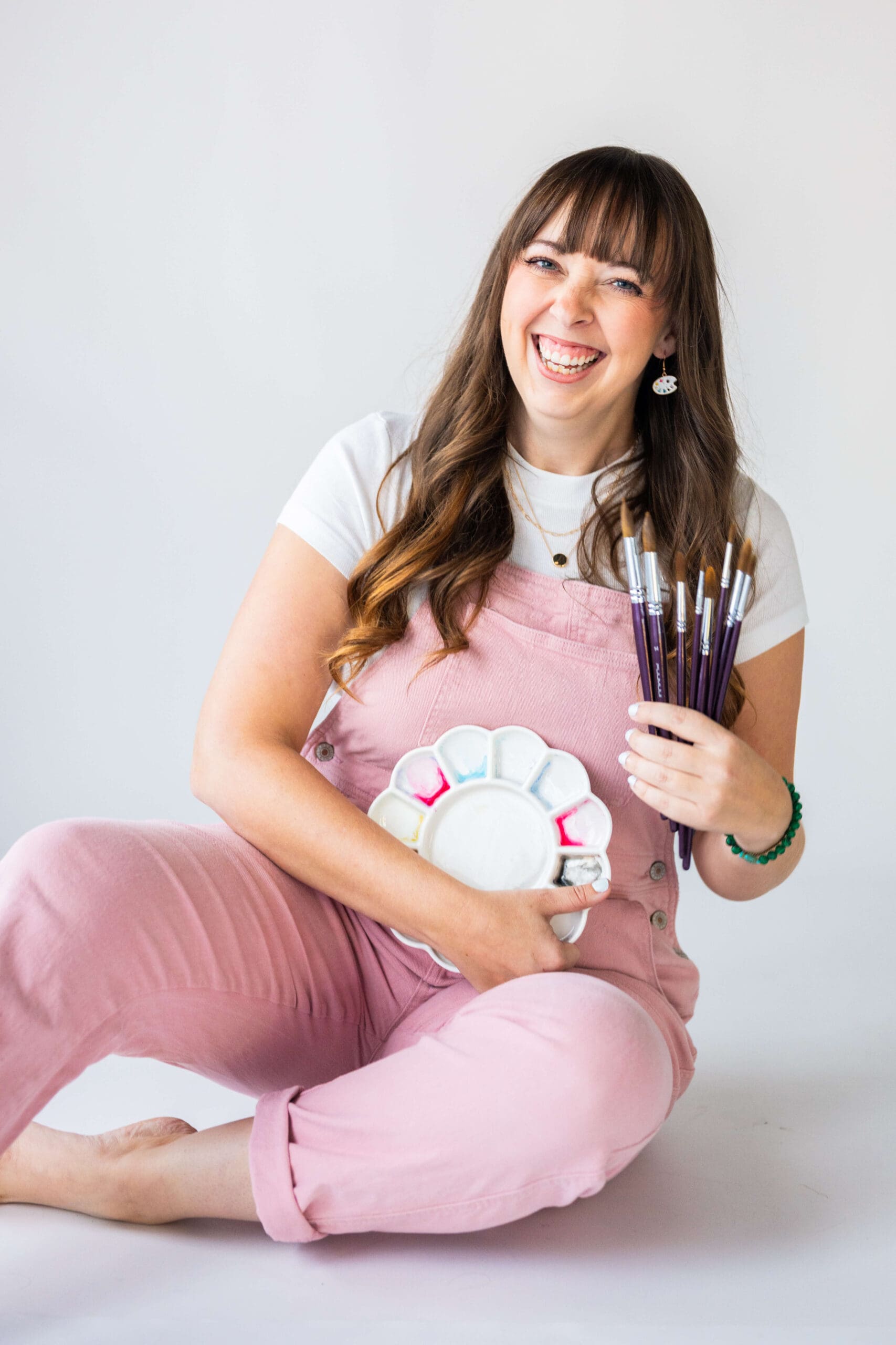 A happy woman in pink overalls sits on the floor holding a painting tray and paintbrushes