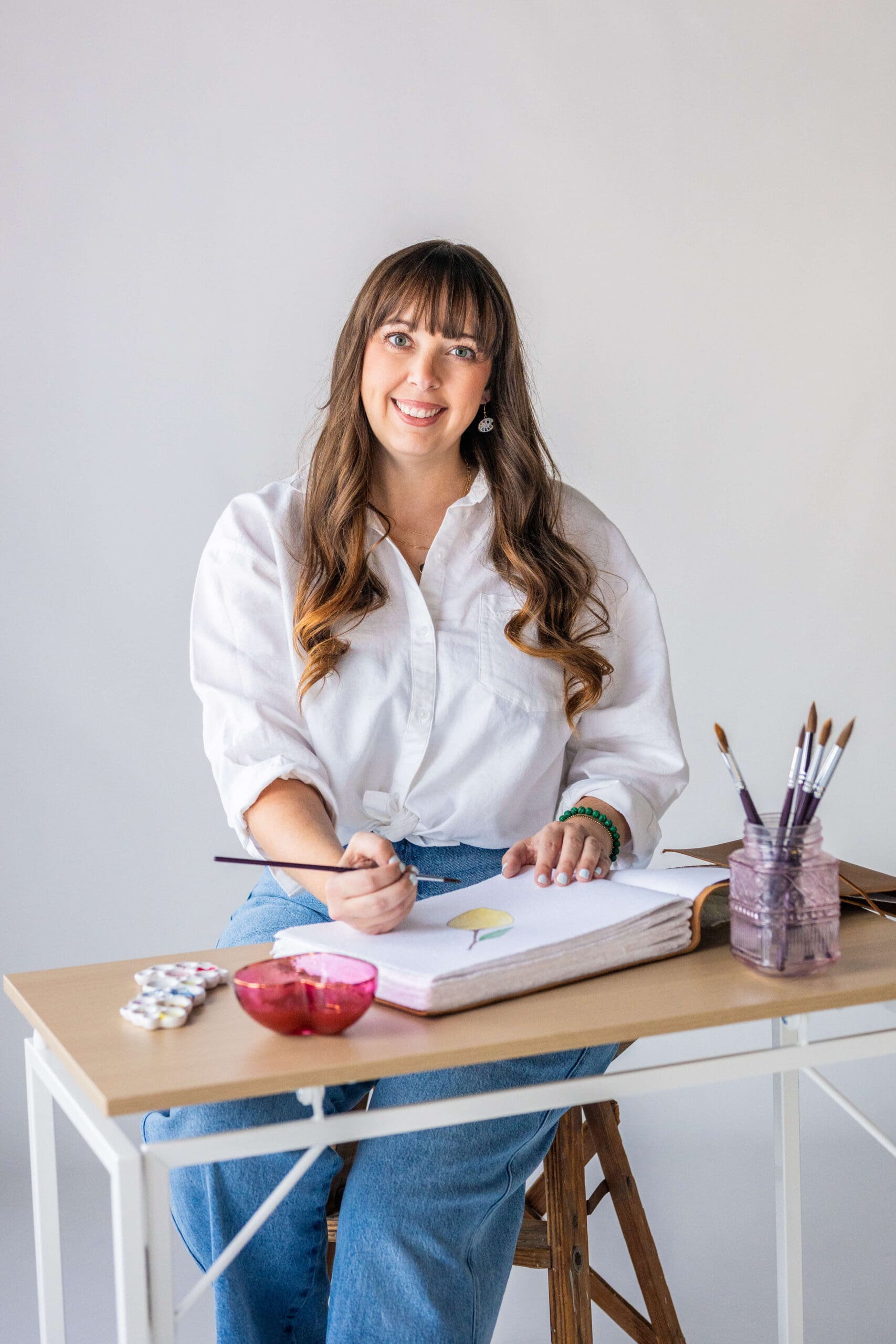 A woman sits at a tall desk painting in a large book