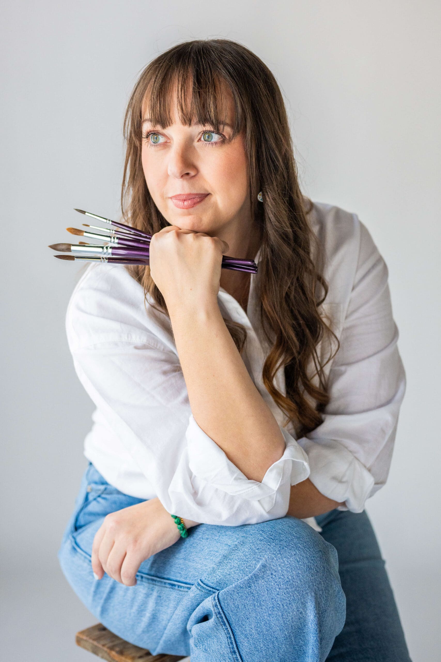 A woman sits on a stool in a white shirt holding a handful of paintbrushes