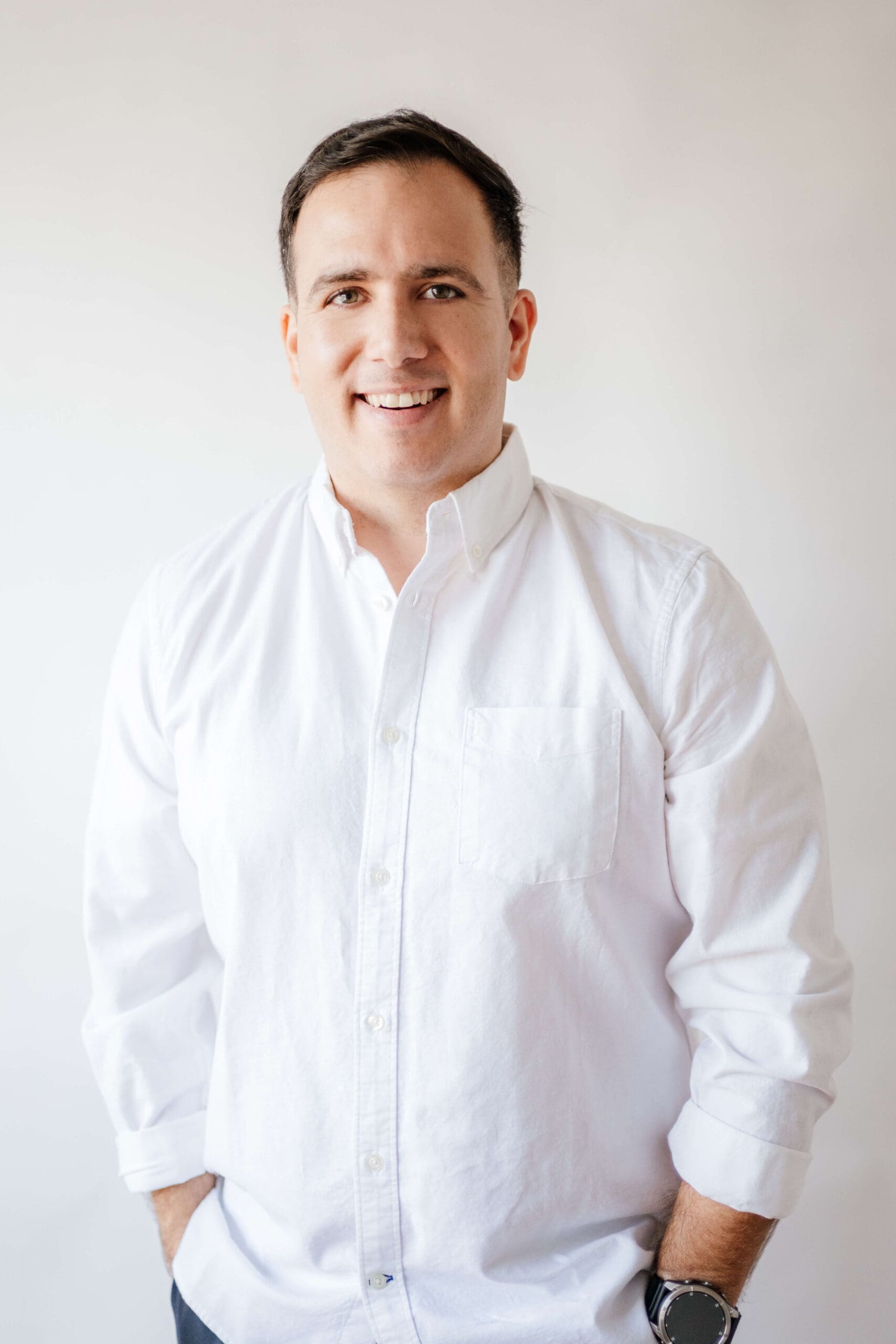 A man in a white shirt smiles while standing in a white room with hands in his pockets