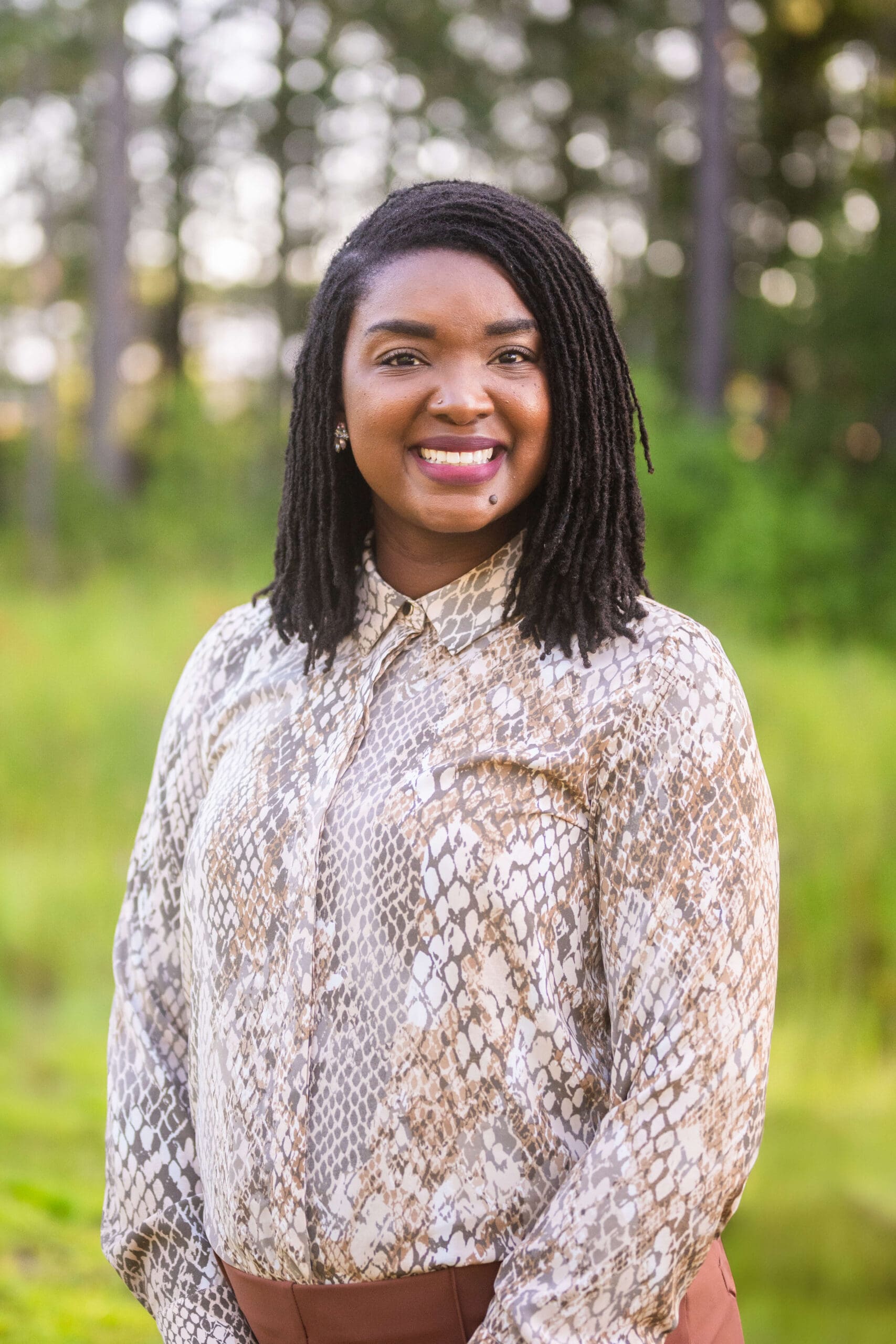 A woman in a pattern print shirt smiles while standing in a forest at sunset during her personal branding headshots