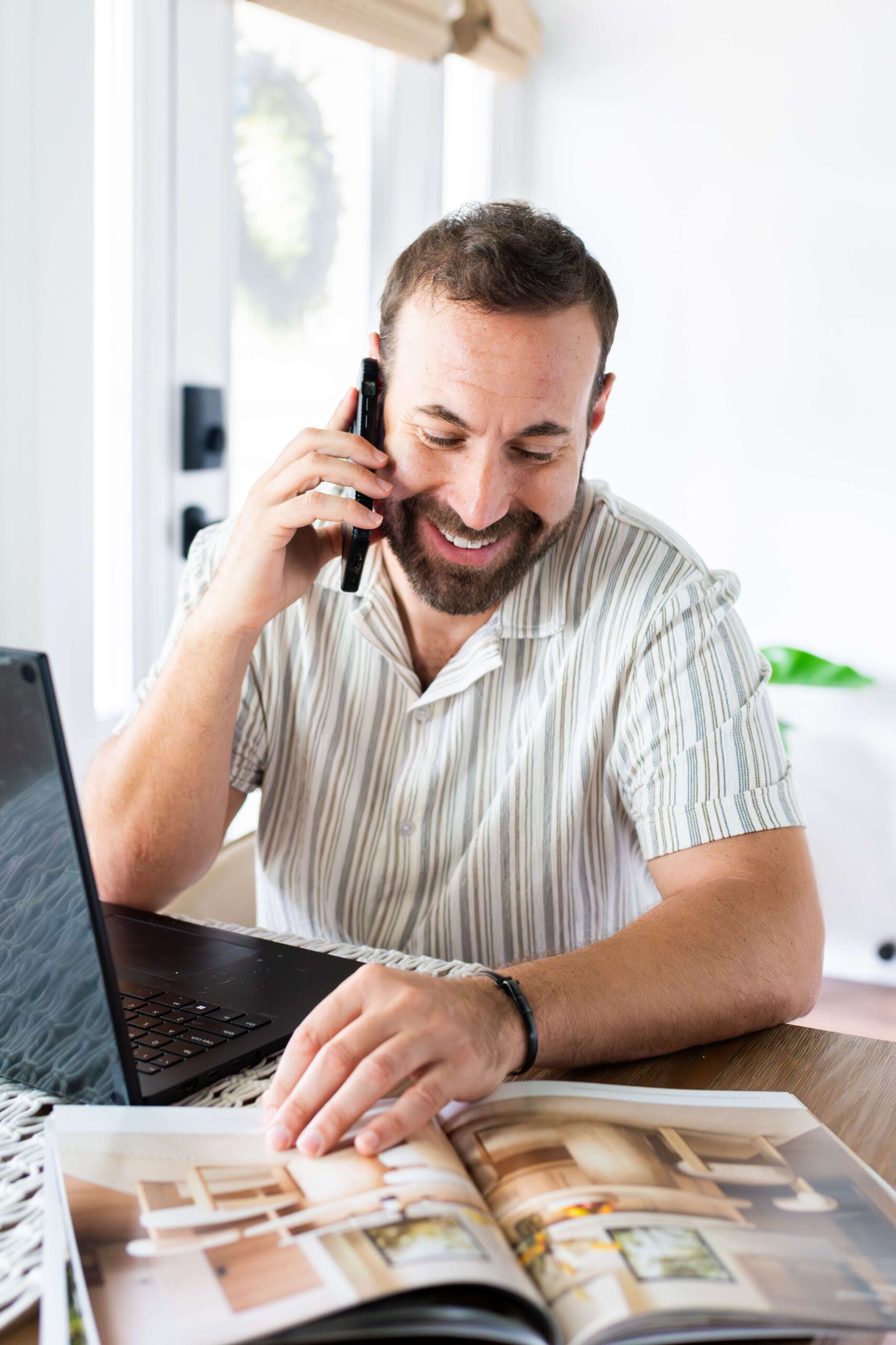 A man sitting at a table on the phone looking through a catalogue at his computer during his personal branding headshots