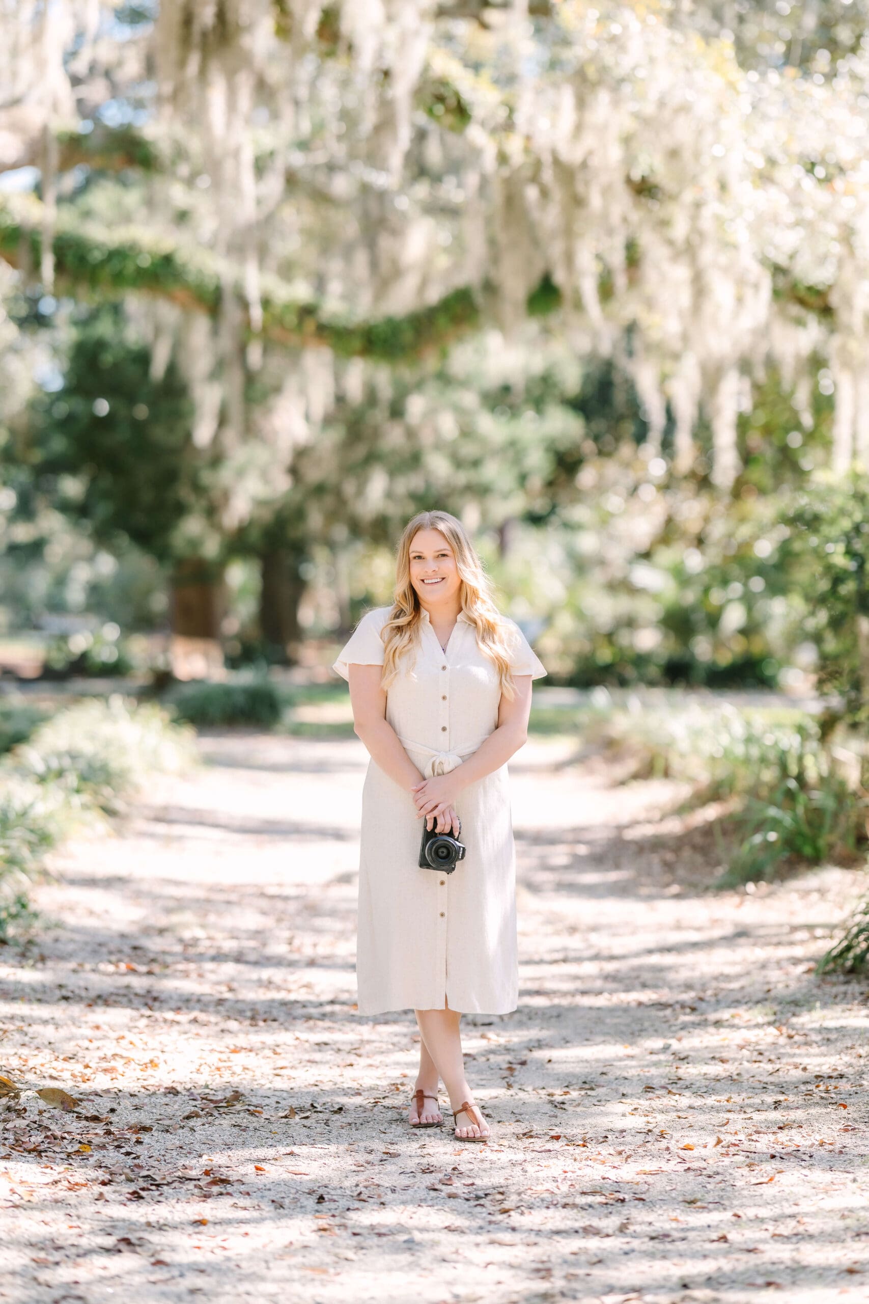 A woman in a creme dress stands in a park trail holding a camera