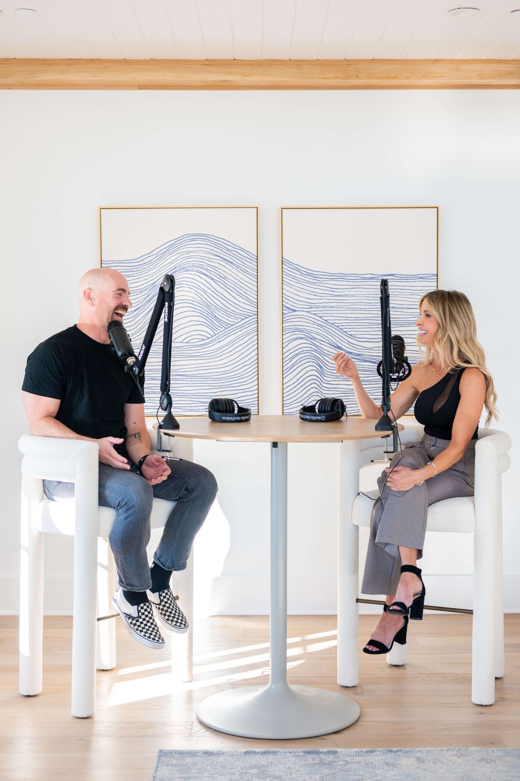 A man and woman in black sit at a high table chatting into microphones for a podcast
