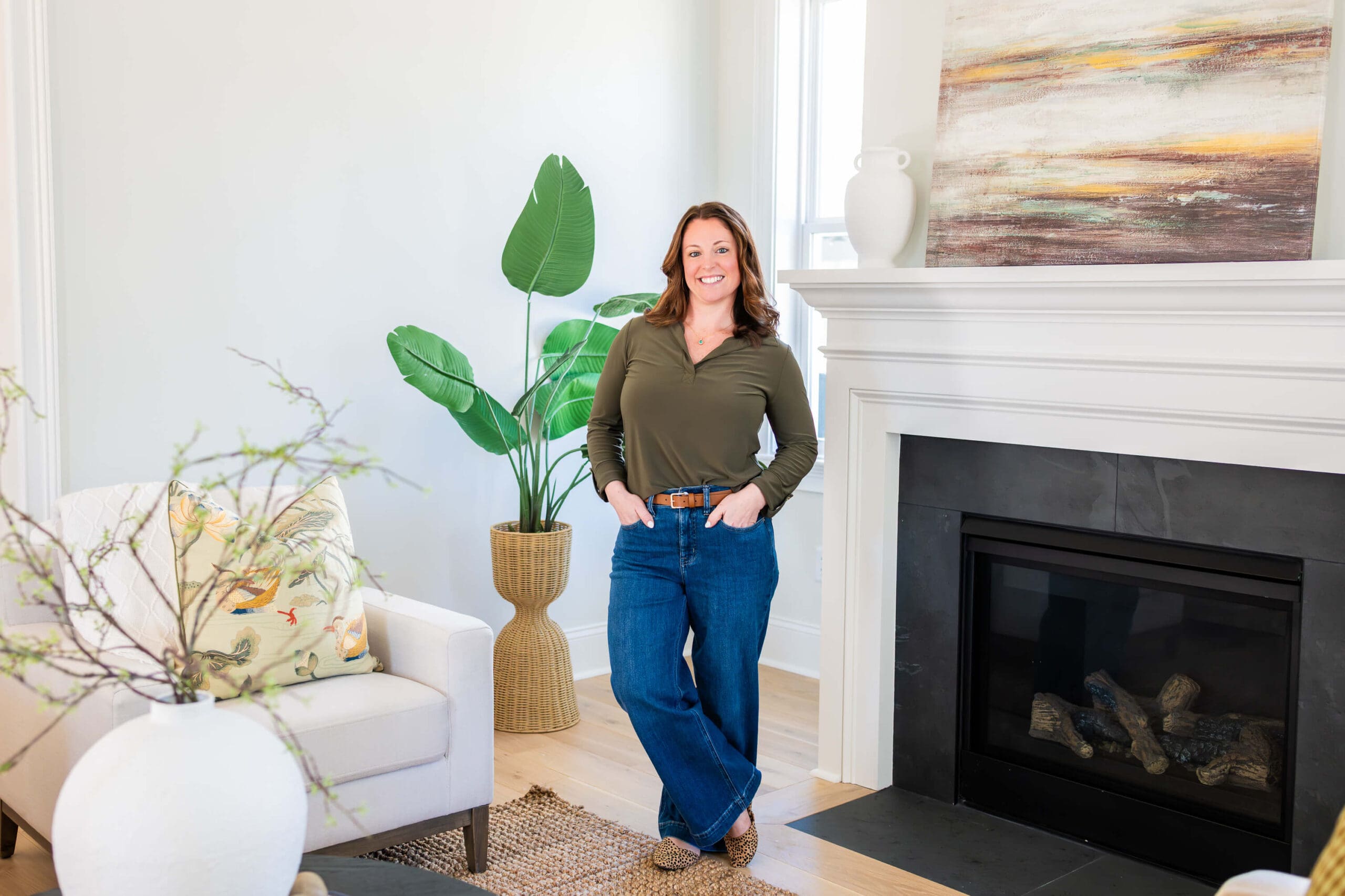 A smiling woman in a green blouse and hands in her jean pockets while standing in a living room