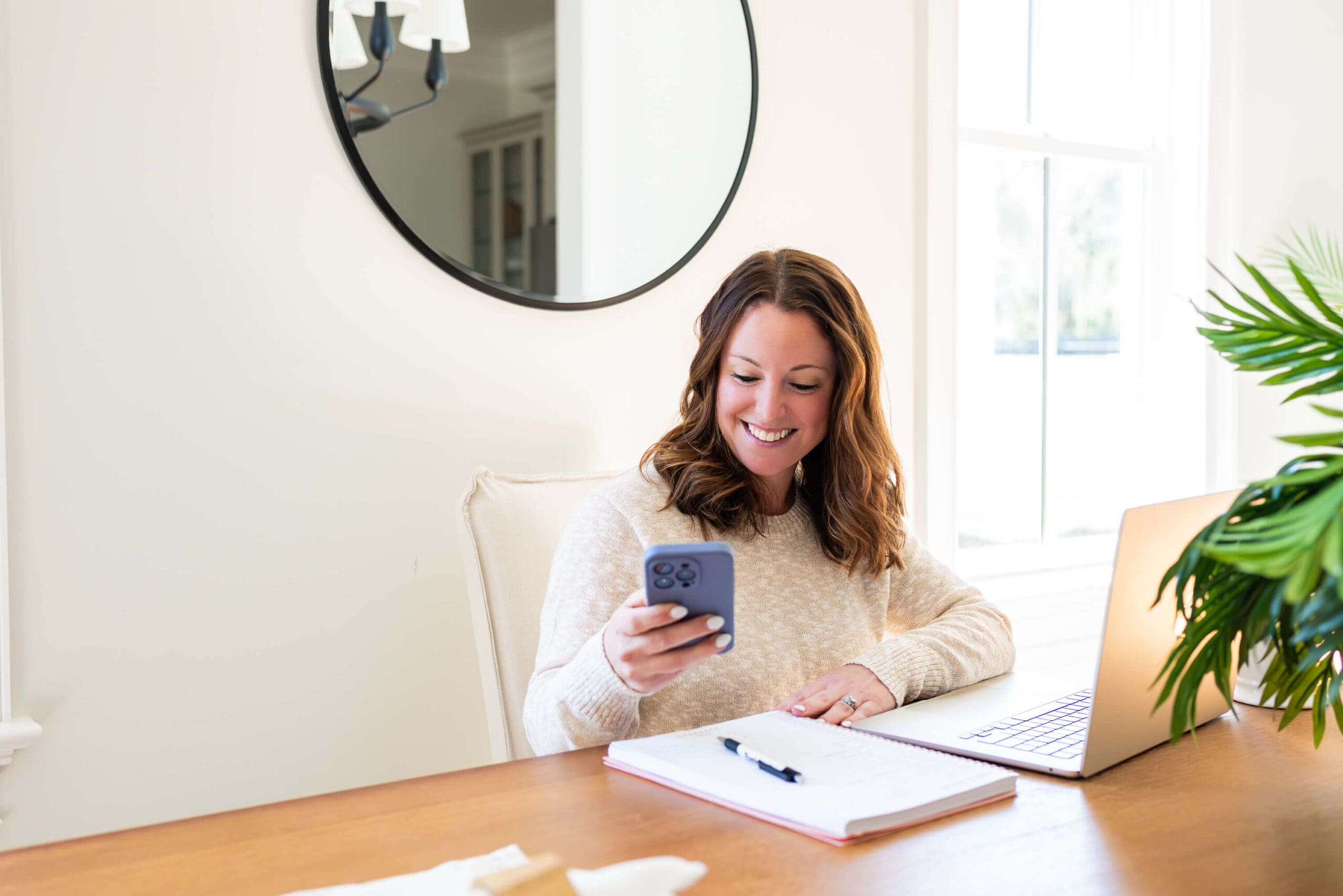 A happy woman checks her phone while working at a dining room table