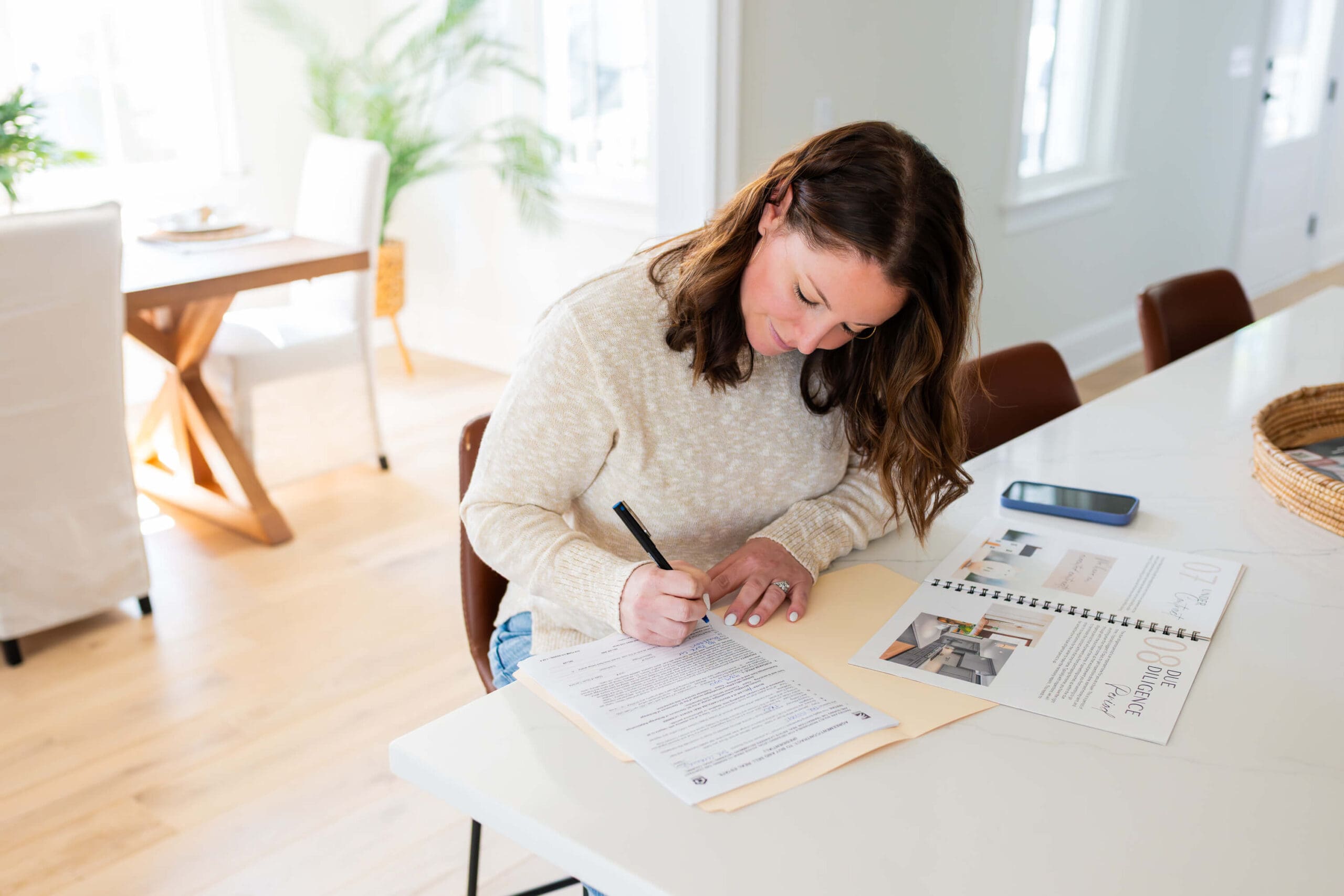 A woman in a cream sweater signs paperwork at a kitchen island after learning How to Stand Out in a Saturated Market