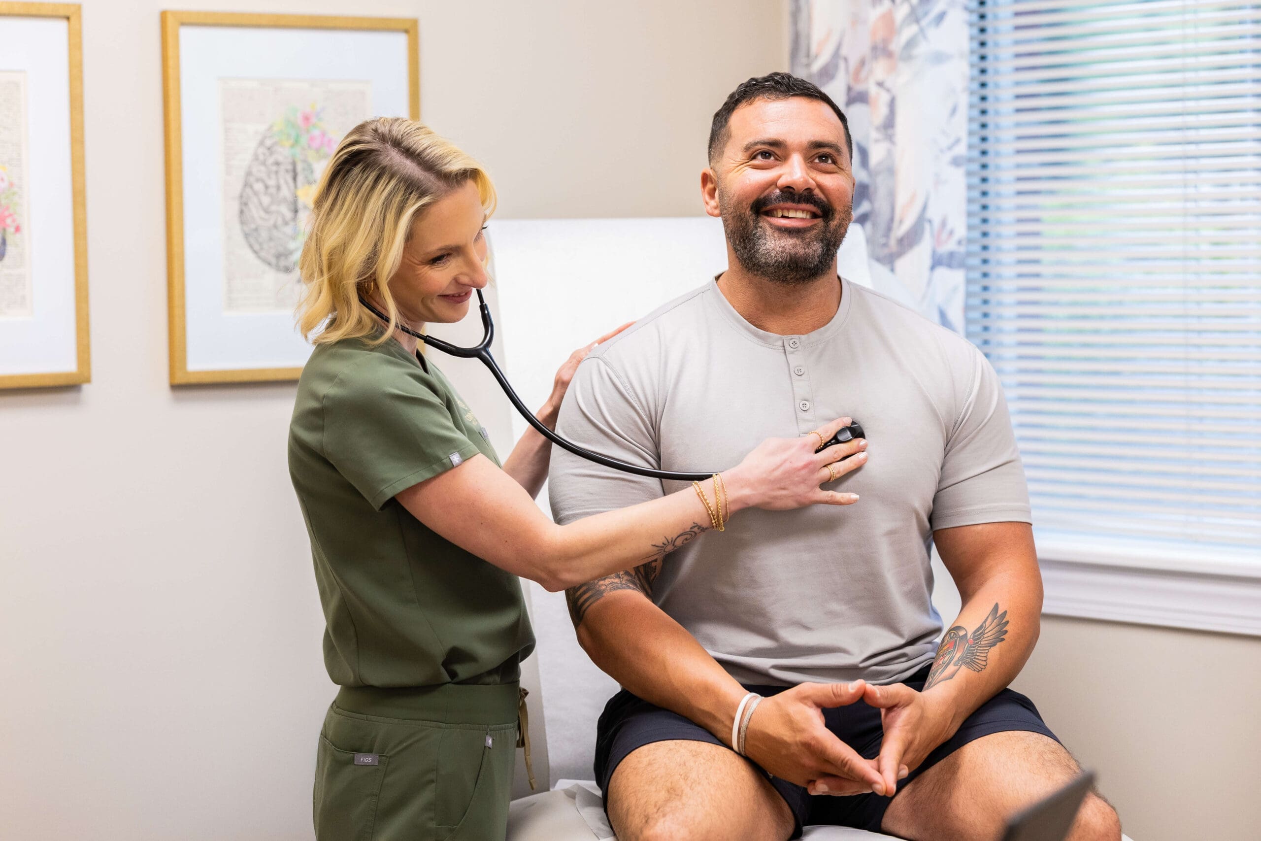 A doctor in sage scrubs listens to her stethoscope on a happy man's chest