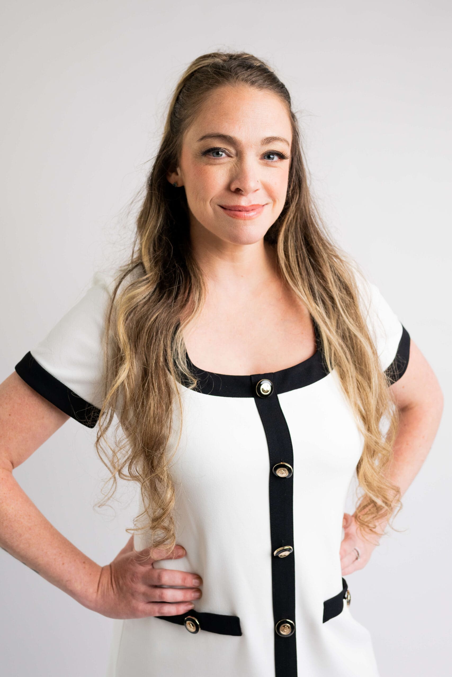 A happy woman in a white and black dress stands with hands on her hips in a studio