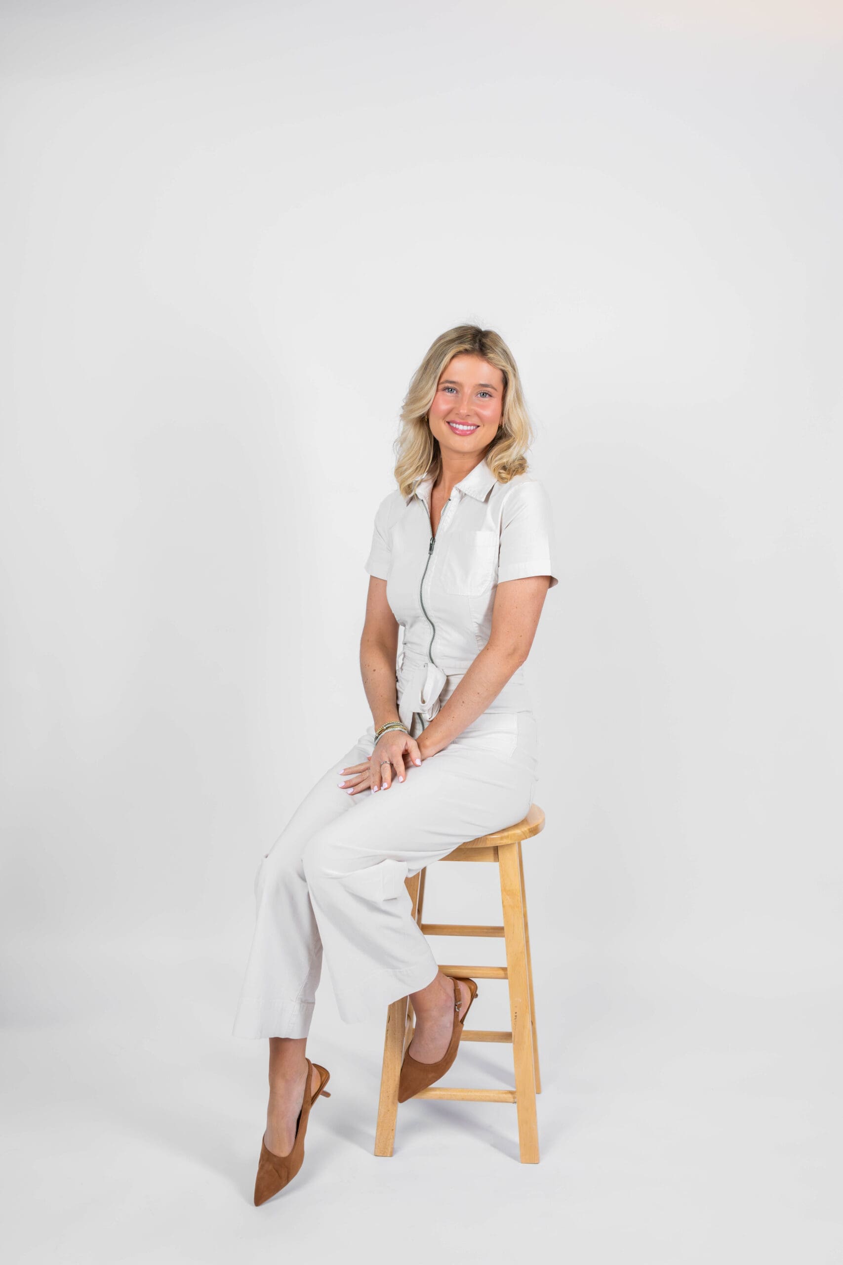 A smiling blonde woman sits on a stool with hands in her lap in a white in a white romper in a studio after finding boutiques in bluffton, sc