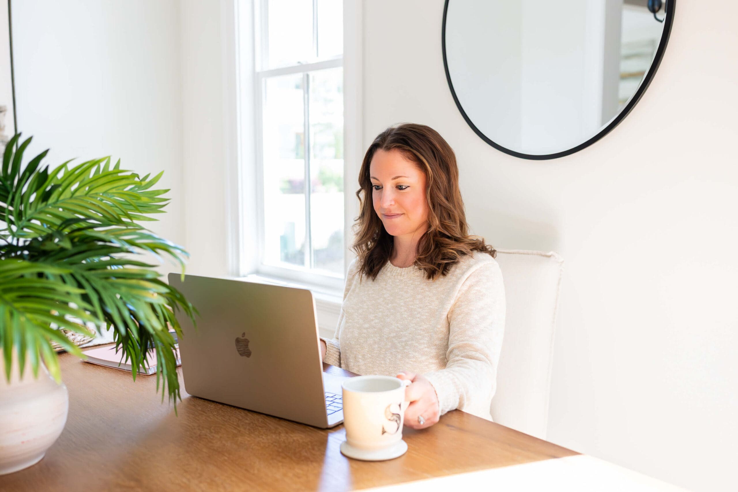A woman smiles while working on a laptop at a kitchen table with a coffee for her real estate branding photos