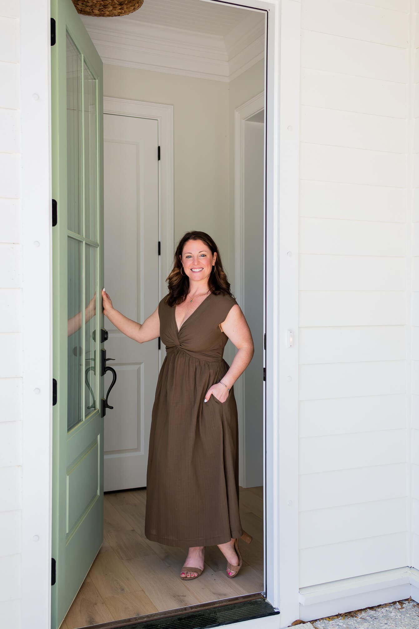 A happy woman stands smiling in a doorway in a brown dress with hand in pocket