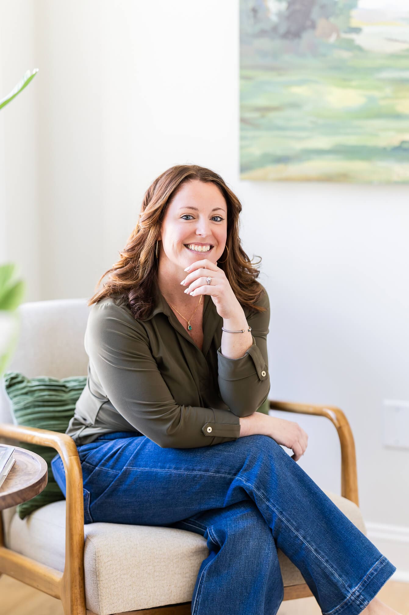 A smiling woman sits in a living room chair in jeans and green blouse leaning on her crossed legs during her real estate branding photos
