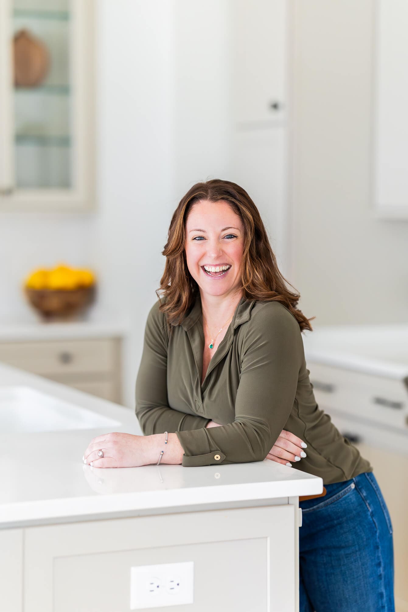 A smiling woman in a green blouse and jeans leans in a whtie kitchen for her real estate branding photos
