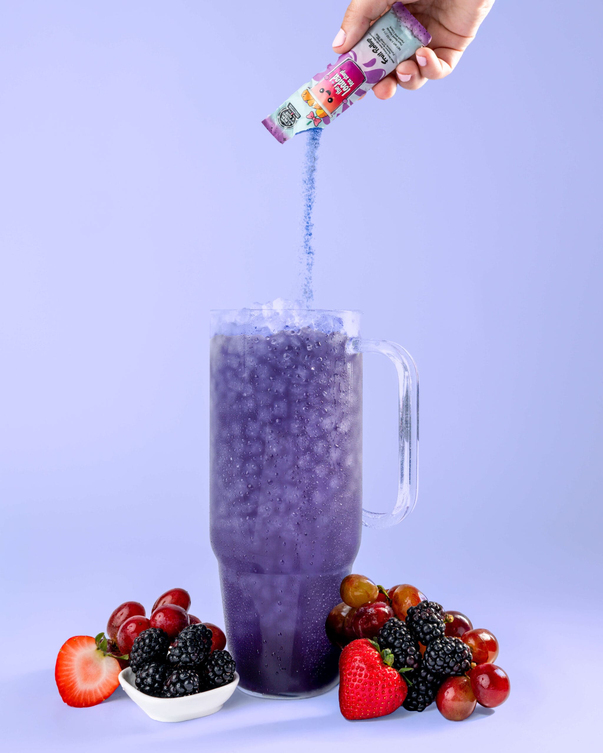 A drink powder is poured into a purple drink in a large clear cup surrounded by berries