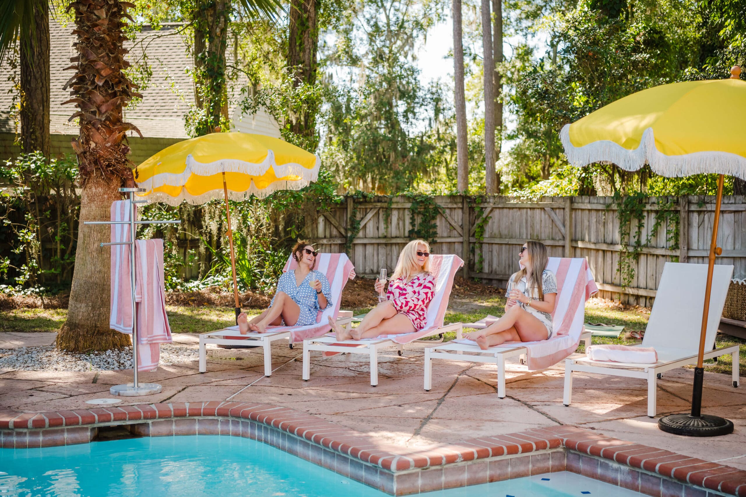 Three women lounge by the pool drinking champagne and laughing